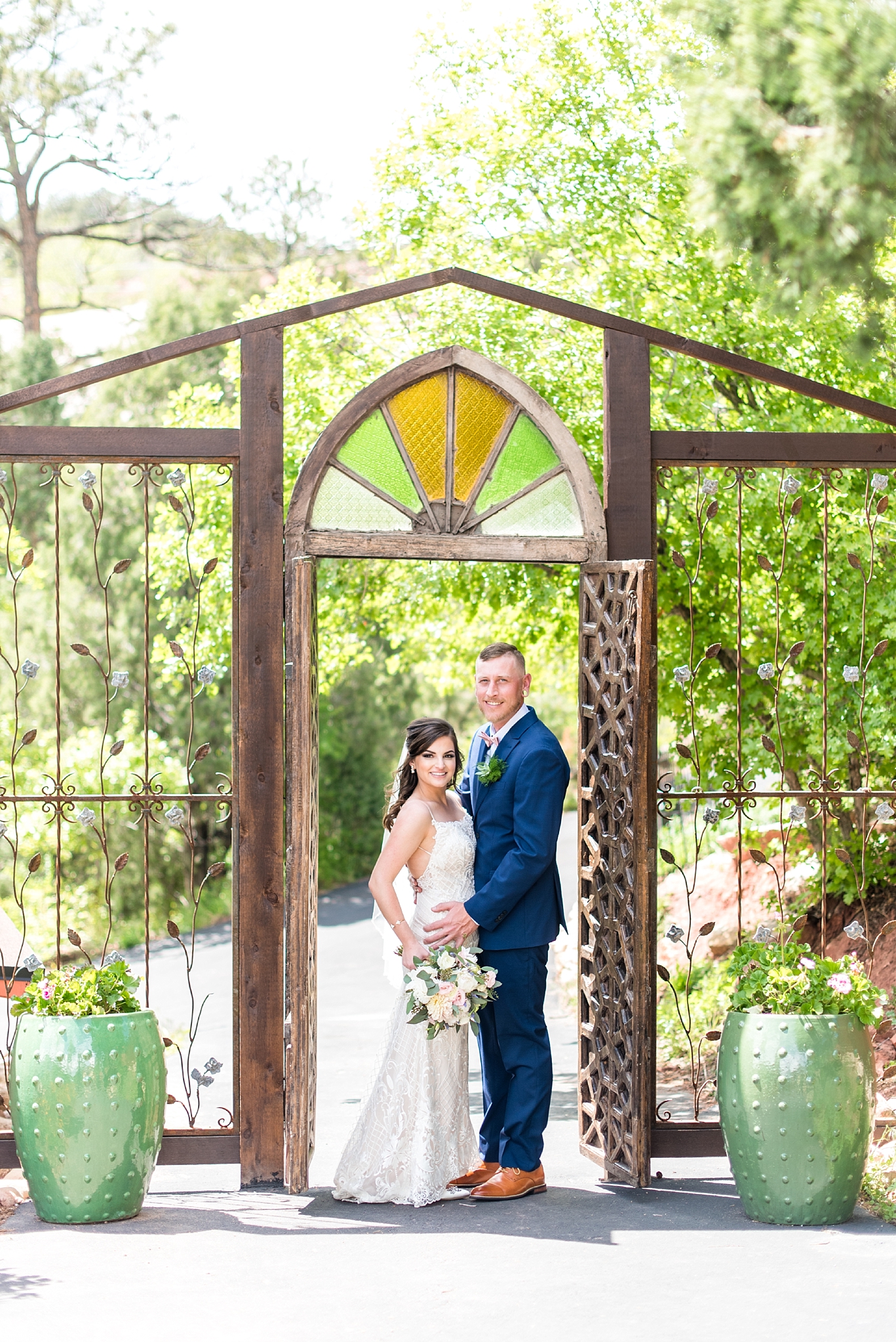 bride and groom standing in a doorway with green and yellow stained gladd over them and wrought iron bars on either side. bride and groom are looking and smiling at the camera. groom is wearing a blue suit and the bride is wearing a white dress and holding a bouquet with white and pink flowers